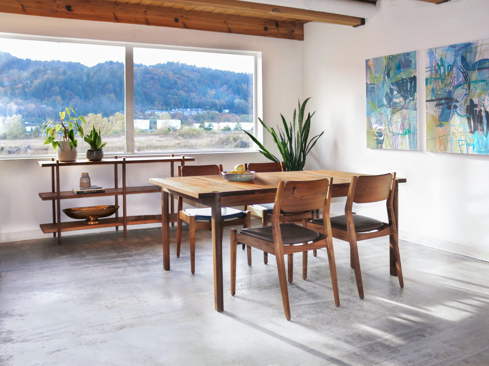 Wood dining table with chairs in a bright room with large window that has a view of forest. 