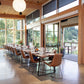 Modern dining area with long table and chairs near large windows with a view of greenery.