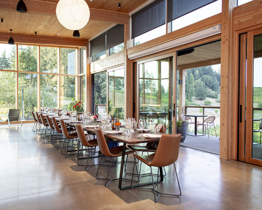Modern dining area with long table and chairs near large windows with a view of greenery.