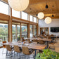 Modern dining area with wooden tables and chairs, large windows, and a view of greenery outside.
