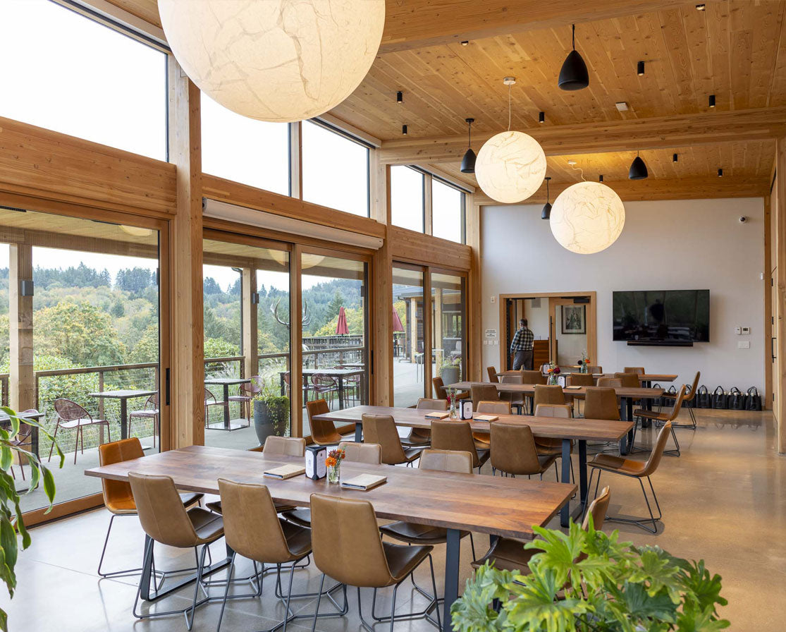 Modern dining area with wooden tables and chairs, large windows, and a view of greenery outside.