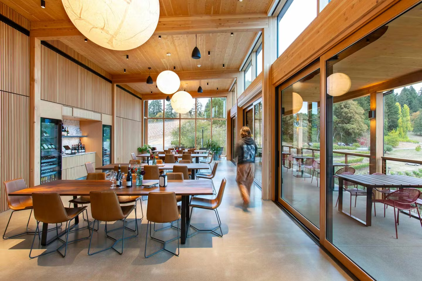 Modern office interior with wooden tables and chairs, large windows, and a person walking.